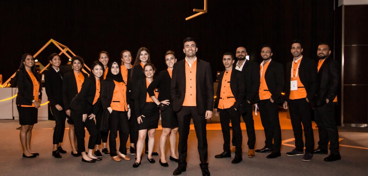 The AladdinB2B team of about sixteen people in black and orange uniform at GITEX, Aladdin Ismail standing centre front